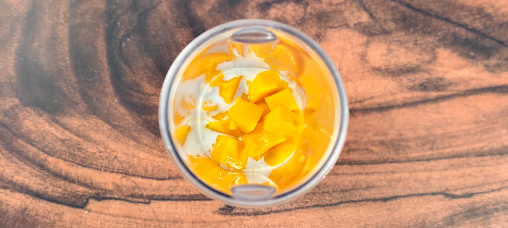 A blender jar filled with cubed fresh mangoes and coconut cream.