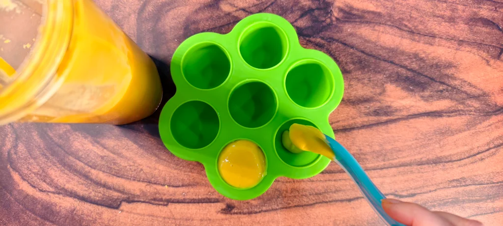 A silicone popsicle mold being filled with mango coconut puree using a spoon.