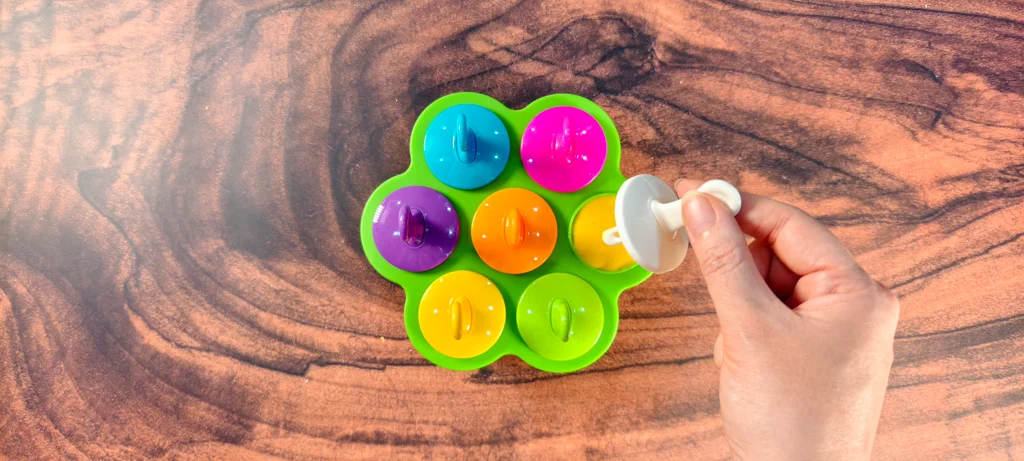 A silicone popsicle mold with popsicle handles inserted into the mango coconut puree. A hand is holding the final popsicle handle to be inserted into the popsicle mold.