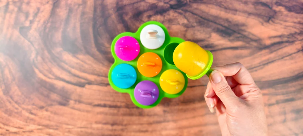 Popsicle handles inserted into a popsicle mold with a hand that is holding a frozen mango coconut popsicle.