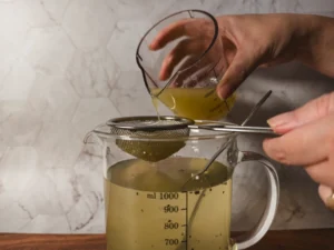 A hand pouring lemon juice from a measuring cup through a strainer into a measuring cup filled with coconut water and chia seeds.